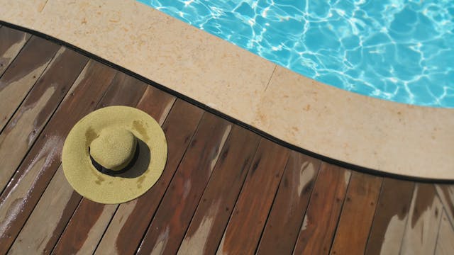 A wide-brimmed straw hat rests on a wet wooden pool deck beside a clear blue swimming pool.